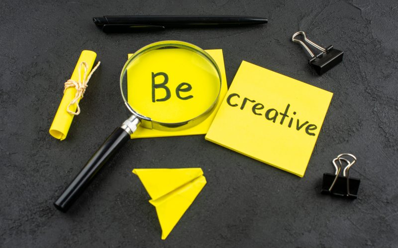 bottom-view-be-creative-written-yellow-sticky-note-lupa-binder-clips-pen-dark-background